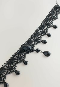 Black lace choker with an intricate web pattern, featuring a central faceted black stone and dangling black bead accents.