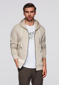 Felpa con zip beige, dotata di lacci, con tasche laterali e zip decorata. Indossata sopra una t-shirt bianca con testo grigio.