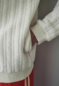 Fehér, textúrált pulóver steppelt mintával, bordázott mandzsettákkal és kenguruzsebbel, piros nadrággal, fehér csíkokkal párosítva.