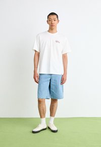 White cotton t-shirt with a small red logo, paired with light blue denim shorts. Worn with white socks and cream shoes with black soles.
