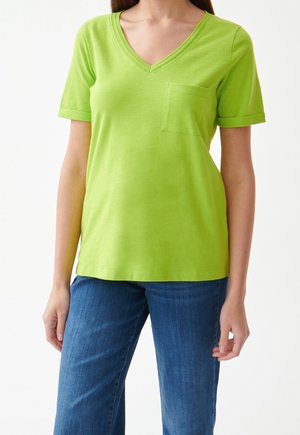 Femme portant un T-shirt vert citron à col en V avec une poche sur la poitrine et un jean en denim bleu, debout devant un fond blanc.