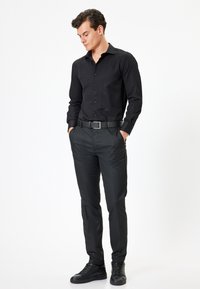 Camicia nera button-up con una texture a coste, abbinata a pantaloni neri slim-fit e scarpe da ginnastica scure. La cintura presenta una fibbia rettangolare in argento.