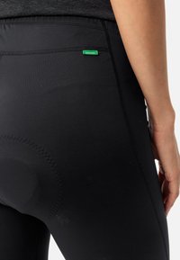 Czarne legginsy wykonane z elastycznego materiału, z płaskim pasem w talii i małą zieloną metką. Zawierają szczegóły w postaci teksturowanych steków.