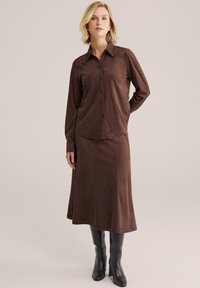 Femeie purtând o cămașă maro cu mâneci lungi și nasturi și o fustă midi asortată, cu cizme negre cu toc până la genunchi, stând în picioare pe un fundal simplu.