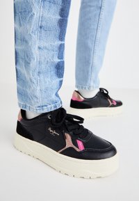 Baskets en cuir noir avec des accents rose et beige, dotées d'une épaisse semelle blanche et de lacets noirs, associées à un jean délavé clair et usé.