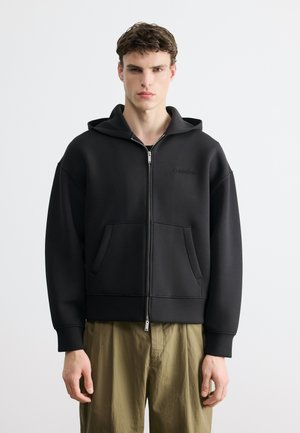 Férfi fekete, cipzáros kapucnis pulóvert visel elülső zsebekkel és olívazöld nadrágot, fehér, egyszínű háttér előtt áll.