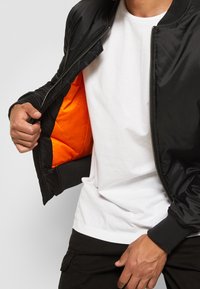 Chaqueta bomber negra con cierre de cremallera y puños acanalados, con un forro naranja brillante. El modelo lleva una camisa blanca debajo.