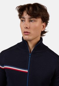 Maglione navy con colletto alto e chiusura con zip. Presenta un tessuto a maglia testurizzato e una striscia orizzontale in rosso, bianco e azzurro sulla parte anteriore.