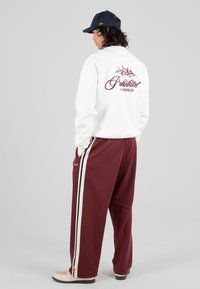 Persona che indossa una felpa bianca con il testo "Proibito 119 Berlino" e pantaloni sportivi bordeaux con strisce laterali bianche e nere, in piedi contro uno sfondo bianco.