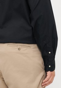 Black long-sleeve shirt with buttoned cuffs, worn with light beige pants featuring a back pocket with a buttoned closure.