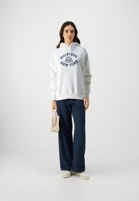Felpa bianca con cappuccio e grafica testuale blu, abbinata a jeans a gamba larga blu e scarpe da ginnastica bianche. Tiene una piccola borsa beige.