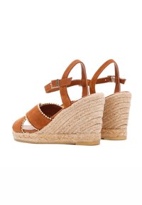 Bruine suede wedge sandalen met een gevlochten jute hak. Kenmerkt zich door kruissandaalbandjes versierd met kleine gouden kralen en een enkelband met gesp.