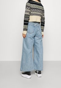 Gekürzter Strickpullover mit geometrischen Mustern in Schwarz, Creme und Grün, kombiniert mit hellblauen, weit geschnittenen Jeans und schwarzen Turnschuhen.