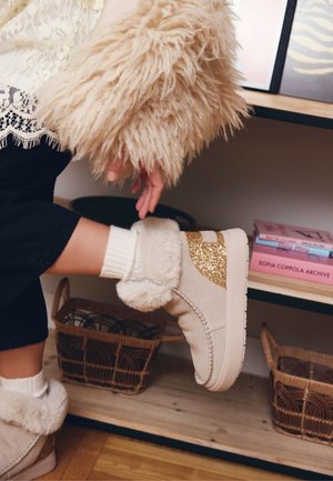 Stivaletti alla caviglia in pelliccia sintetica beige con dettagli glitterati, fodera testurizzata e suola piatta. Indossati con calzini bianchi lavorati a maglia e abbinati a pantaloni scuri.