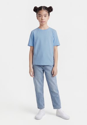 Νεαρό άτομο όρθιο, φοράει ανοιχτό μπλε μπλουζάκι, ανοιχτό μπλε τζιν, λευκά αθλητικά παπούτσια, με δύο κοτσίδες στο πλάι, ουδέτερη έκφραση.