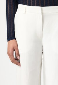 Pantaloni bianchi di lino con una texture liscia, dotati di vita piatta e tasche laterali discrete, abbinati a un maglione blu navy a costine.