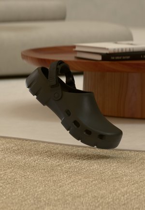 Zoccolo nero in gomma con cinturino regolabile che fluttua sopra un tappeto beige strutturato, tavolino rotondo in legno e divano sfocati sullo sfondo.