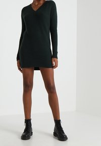 Vestido suéter verde de punto con escote en V y mangas largas, que presenta un patrón texturizado y una silueta semiestructurada, combinado con botas negras.