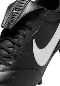 Fekete futballcipő texturált bőrrel, fehér Nike Swoosh-szal és fekete fűzőkkel. párnázott nyelvvel és tüskékkel a tapadás érdekében.