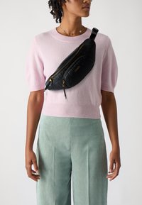 Schwarze gesteppte Bauchtasche mit goldenen Reißverschlüssen, getragen über einem hellrosa kurzärmeligen Pullover, kombiniert mit grün gestreiften Hosen.