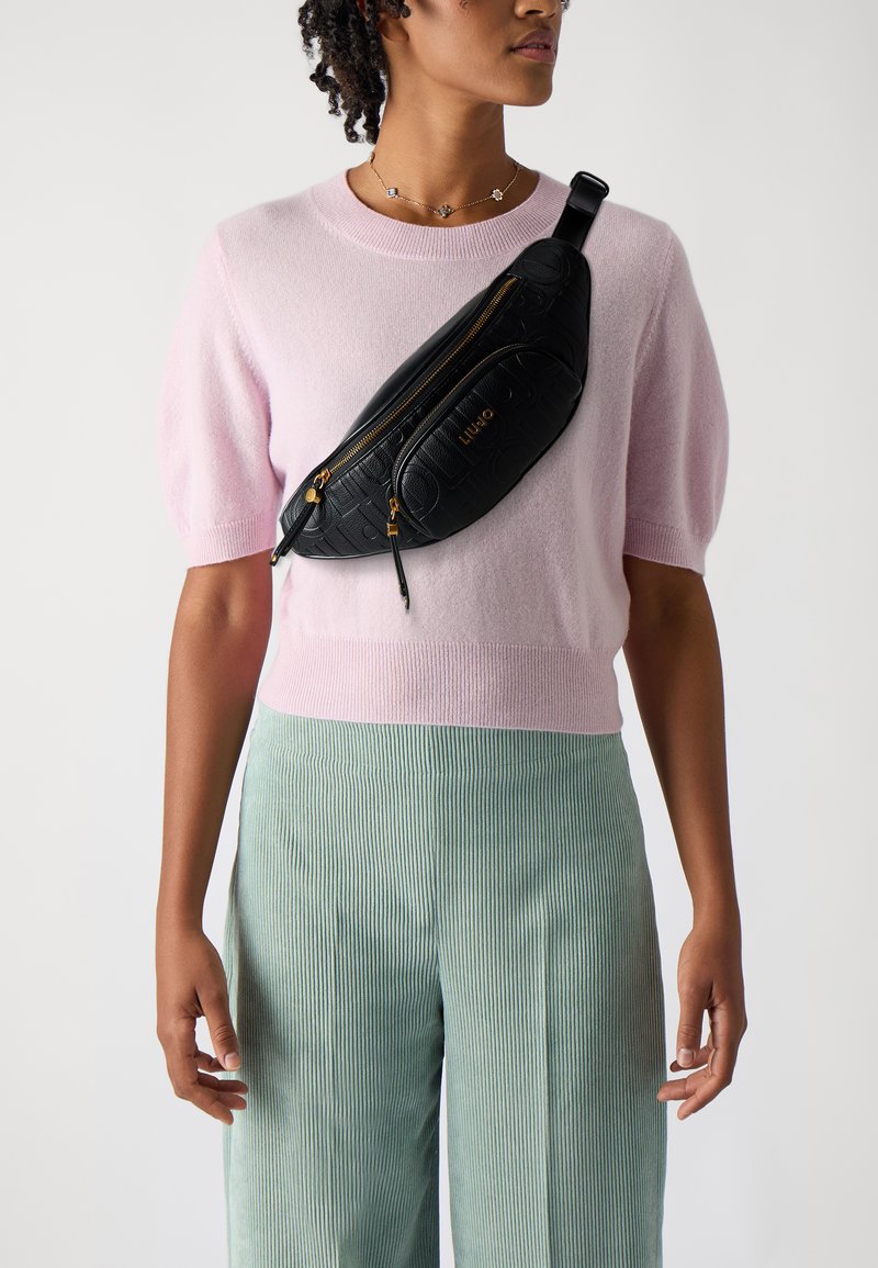 Schwarze gesteppte Bauchtasche mit goldenen Reißverschlüssen, getragen über einem hellrosa kurzärmeligen Pullover, kombiniert mit grün gestreiften Hosen.