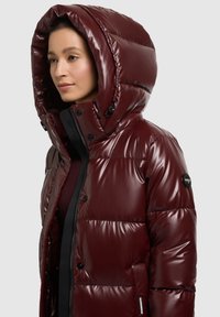 Bordó, fényes felületű pehelydzseki nagy kapucnival, patentos gombokkal és karcsú kialakítással; az ujjon logóval ellátott folttal.