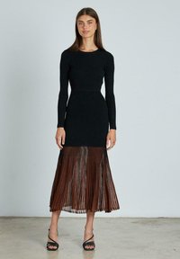 Abito lungo a maniche lunghe di colore nero con sovrapposizione trasparente plissettata marrone, caratterizzato da un corpetto aderente e una gonna fluida. Tacchi neri con cinturini accentuano il look.