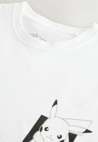 T-shirt blanc avec un graphique de Pikachu partiellement visible près du col, montrant le visage et les oreilles de Pikachu en noir et blanc.