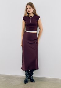 Bordeaux crop top met witte afwerking, gecombineerd met een bijpassende lange rok. Het model draagt kniehoge zwarte leren laarzen. Soepele, aangesloten stof.