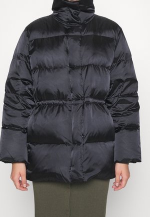 Schwarze Steppjacke mit hohem Kragen und tailliertem Schnitt, getragen über einem olivgrünen, geknöpften Rock, gezeigt von den Schultern bis zur Mitte des Oberschenkels.