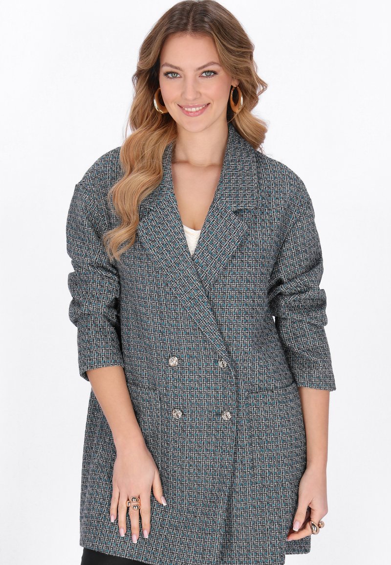 Giovane donna con lunghi capelli mossi che indossa un blazer grigio-blu a doppio petto con texture, orecchini a cerchio e anelli decorativi, sorridente verso la fotocamera.