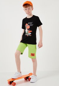 Garçon portant une casquette orange, un t-shirt noir avec un dinosaure sur un skateboard et un short "Ride On !" se tient avec un pied sur un skateboard orange.