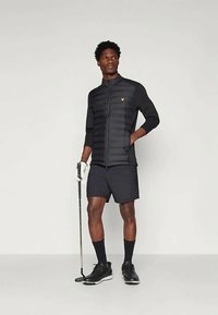 Mann in schwarzer Golfbekleidung mit gepolsterter Jacke, Shorts und Handschuh, der einen Golf-Putter hält und vor einem schlichten, hellen Hintergrund steht.