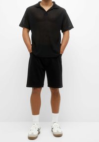 Camisa polo negra de manga corta con tejido texturizado y cuello abierto, combinada con pantalones cortos negros y zapatillas blancas con detalles en color marrón.