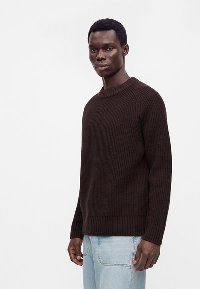 Homme portant un pull en maille marron foncé et un jean bleu clair, debout devant un fond blanc uni.