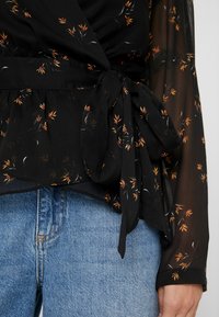 Schwarze, transparente Wickelbluse mit floralen Mustern in Orange und Weiß, ausgestattet mit einem gebundenen Taillen-Detail und langen Ärmeln. Kombiniert mit hellblauen Jeans.