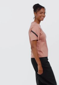 Camiseta de manga corta rosa claro con escote redondeado y un detalle negro en el hombro izquierdo, combinada con pantalones negros. Textura de tela suave.