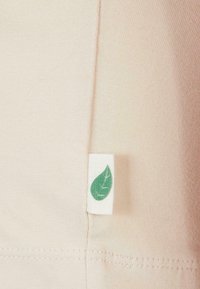 Helles beige Gewebe mit einer glatten Textur, das an der Naht ein kleines, weißes Etikett mit einem grünen Blätterdesign aufweist. Einfaches und minimalistisches Design.