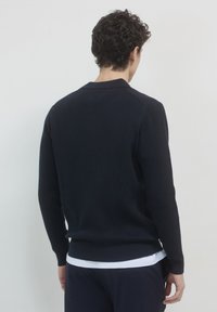 Pull en tricot côtelé bleu marine avec des manches longues et un col ras du cou, présentant une finition texturée et un ourlet droit. Porté par-dessus une chemise blanche.