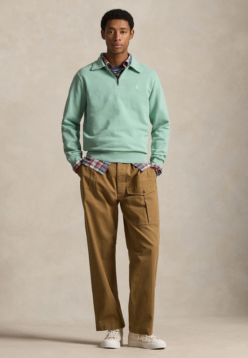 Young man wearing a mint green quarter-zip sweatshirt, plaid shirt, brown cargo pants, and white sneakers, standing with hands in pockets.