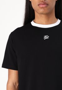 Svart t-shirt med vit krage och en liten broderad logotyp i vitt. Har korta ärmar och en mjuk, slät textur.