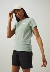 Tricou din bumbac de culoare verde deschis, cu mâneci scurte, buzunar aplicat și branding subtil. Asortat cu pantaloni scurți cargo negri, care au multiple buzunare.