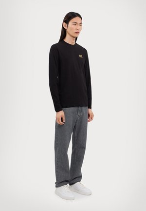 Langermet svart skjorte med en liten gulllogo, kombinert med lysegrå jeans og hvite sneakers, båret av en stående modell.