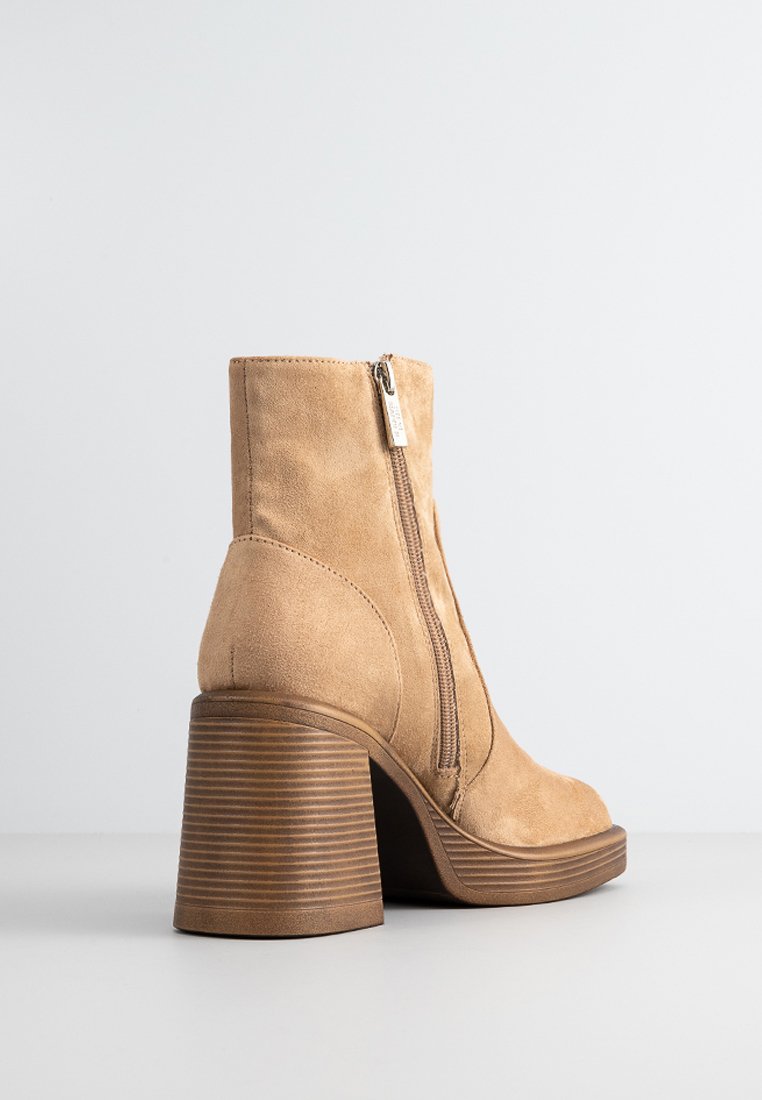 Bottine en suede beige avec un bout rond, un talon en bois empilé et une fermeture éclair sur le côté. Présente une texture lisse et un design épuré.