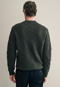 Grober Strickpullover in Dunkelgrün mit einem texturierten Zickzackmuster; gerippter Kragen und Bündchen, lockere Passform und leicht verkürzter Rückensaum.
