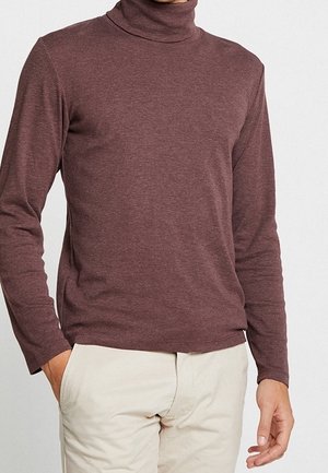 Mann trägt einen enganliegenden braunen Rollkragenpullover und hellbeige Hosen, eine Hand in der Tasche, vor einem einfarbigen Hintergrund.