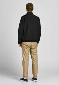 Zwarte lichtgewicht jas met een kraag, elastische manchetten en zoom, gecombineerd met beige broeken en zwarte sneakers met witte zolen.