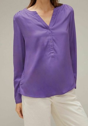 Mujer con blusa morada de manga larga, escote en V y cuatro botones, combinada con pantalones color crema, sobre un fondo neutro.