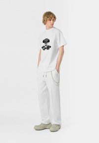 Camiseta blanca de algodón con diseño gráfico negro, combinada con pantalones blancos de cuerda con un acento de cadena y calzado texturizado de color verde claro.