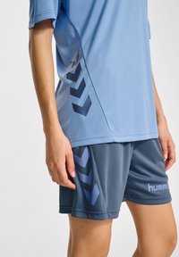 Persoon in een lichtblauw sportshirt en donkerblauwe shorts met chevronpatronen en het "hummel" logo op de shorts.